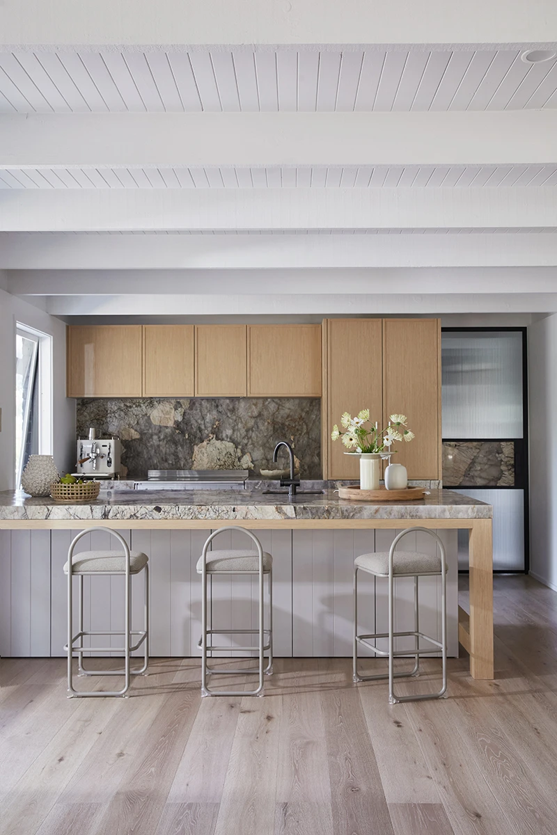Modern kitchen with wood cabinets, marble countertops, three bar stools, and white beamed ceiling on light wood flooring.