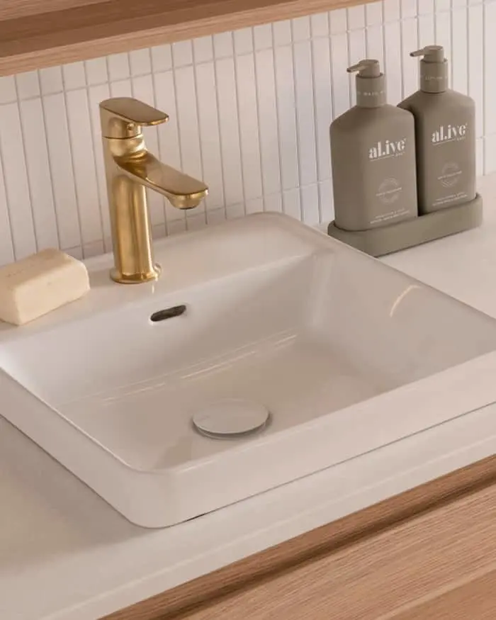 Modern bathroom sink with gold faucet, white basin, soap bar, and two gray pump bottles against white tile backsplash.