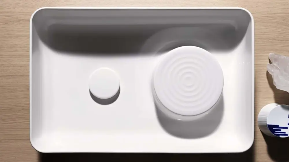 White ceramic tray containing a small circular object and a larger cylindrical container with concentric rings on top.