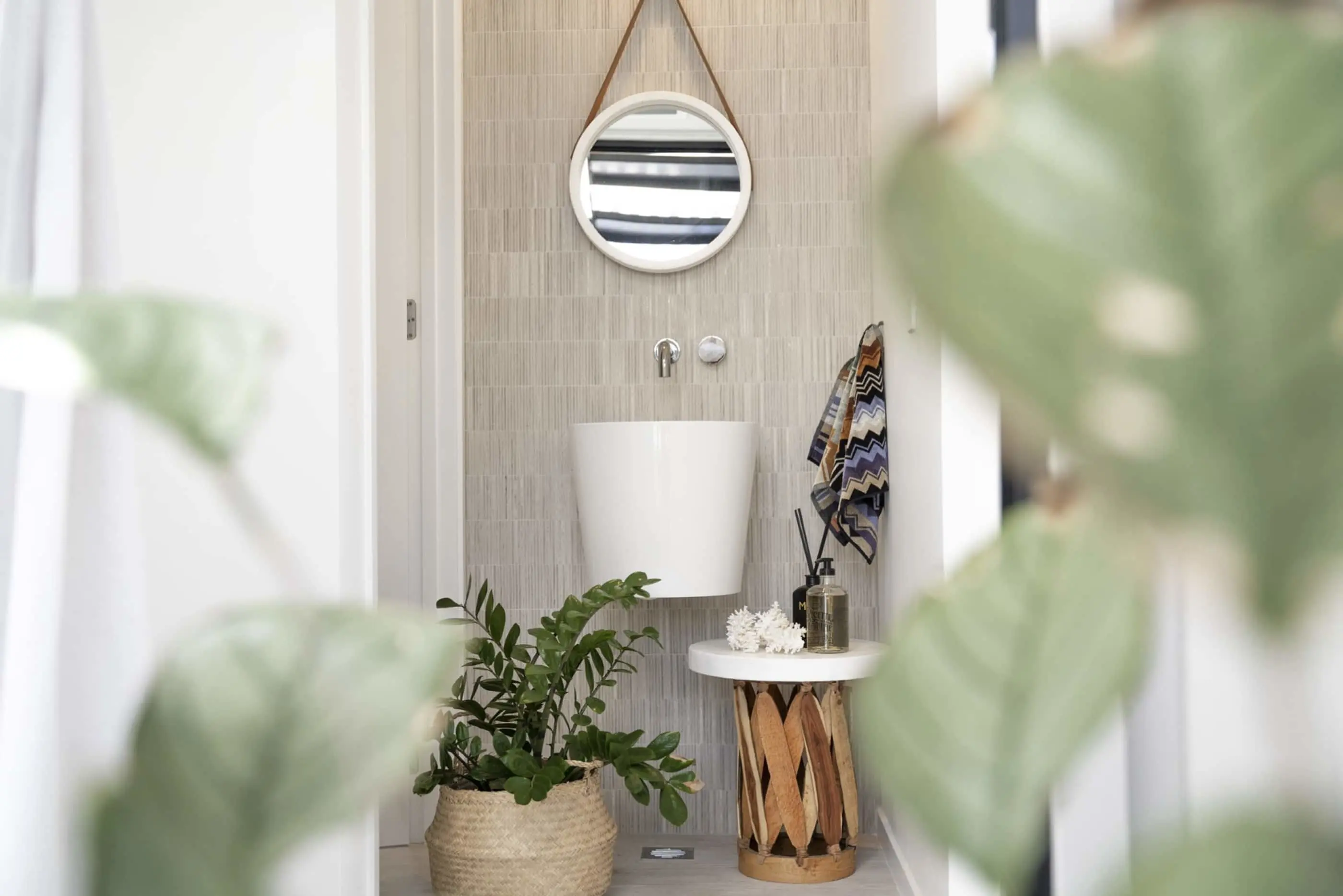 Modern bathroom with white pedestal sink, round mirror, wooden side table, patterned towel, and potted plant in basket.