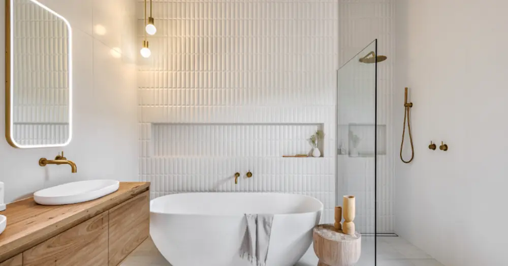 Modern white bathroom with freestanding tub, wooden vanity, gold fixtures, and textured tile walls.
