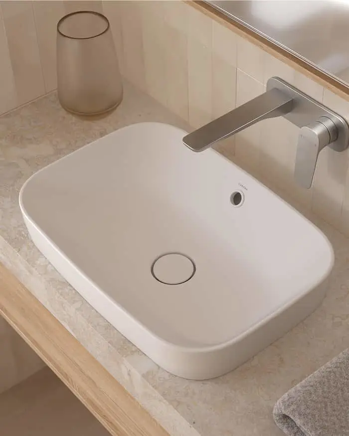 White rectangular bathroom sink on beige countertop with wall-mounted brushed nickel faucet and glass tumbler