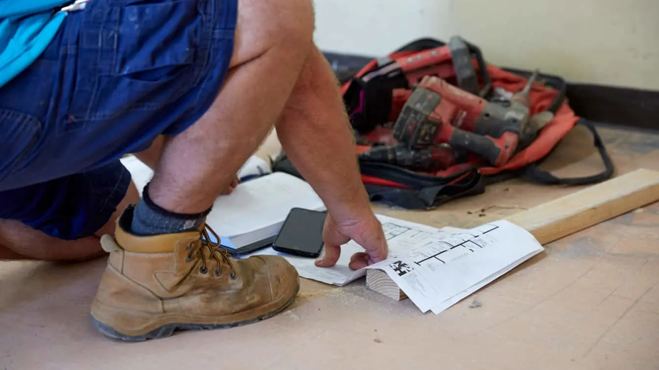 Worker in blue overalls reviewing blueprints on wooden floor with power tools in red bag nearby.