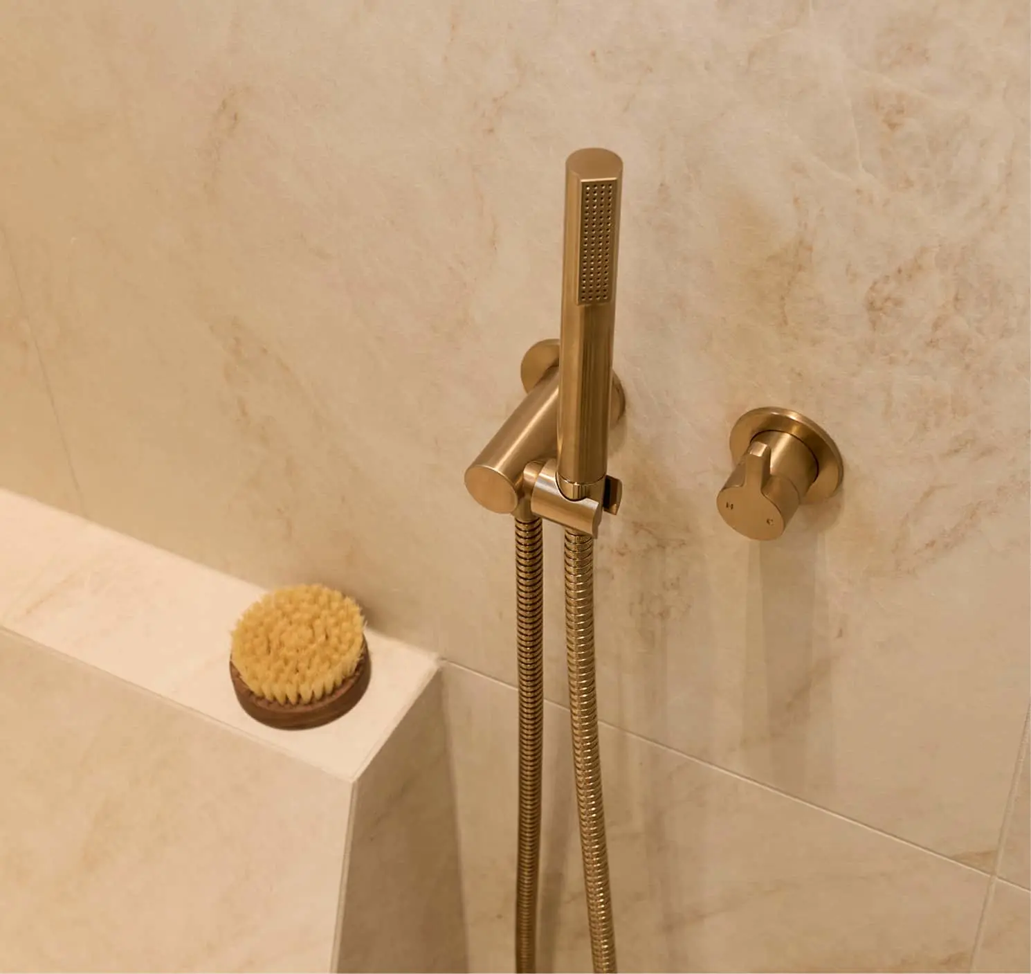 Gold handheld shower head with hose mounted on beige marble wall next to bathtub with natural bristle brush