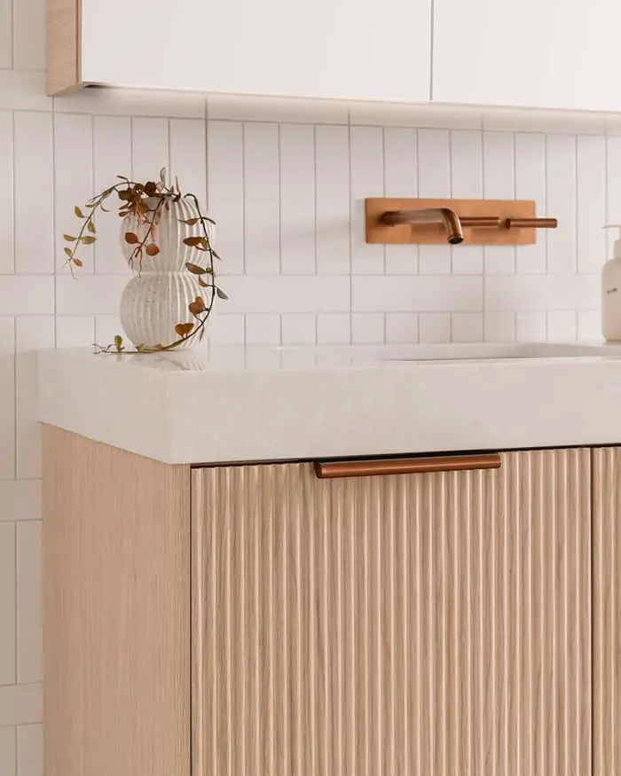 Modern bathroom vanity with white countertop, wooden cabinet, copper faucet, and white vase with dried plant.