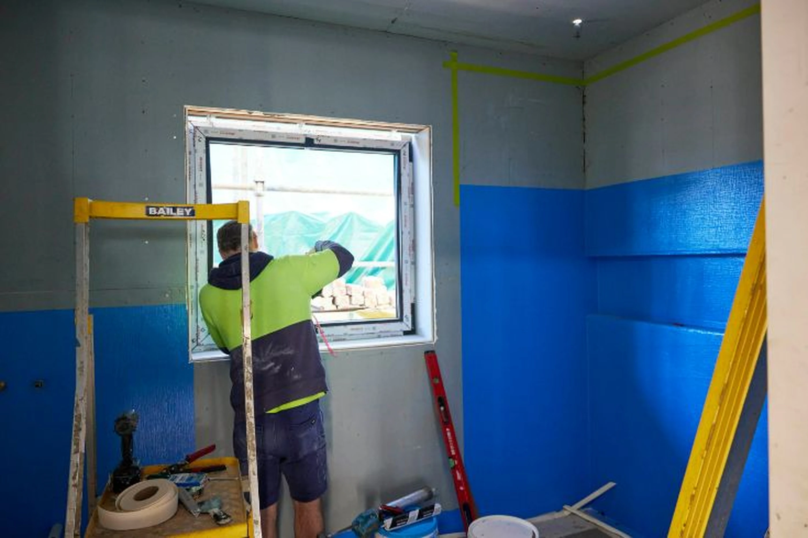 Construction worker installing a window in a room with partially painted blue walls and yellow scaffolding.