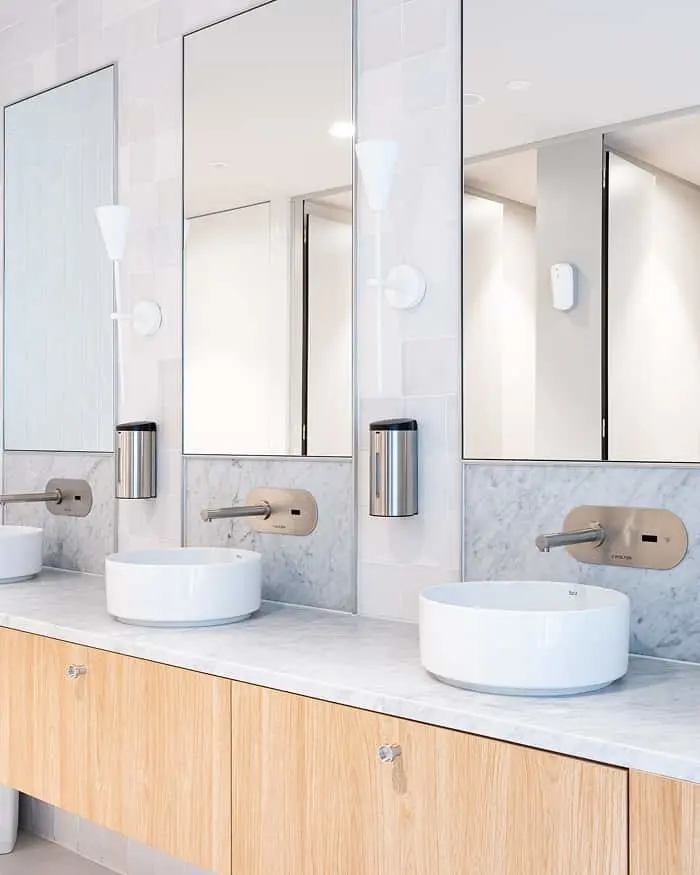 Modern bathroom with two white vessel sinks, wall-mounted faucets, large mirrors, and light wood cabinetry on marble countertop.