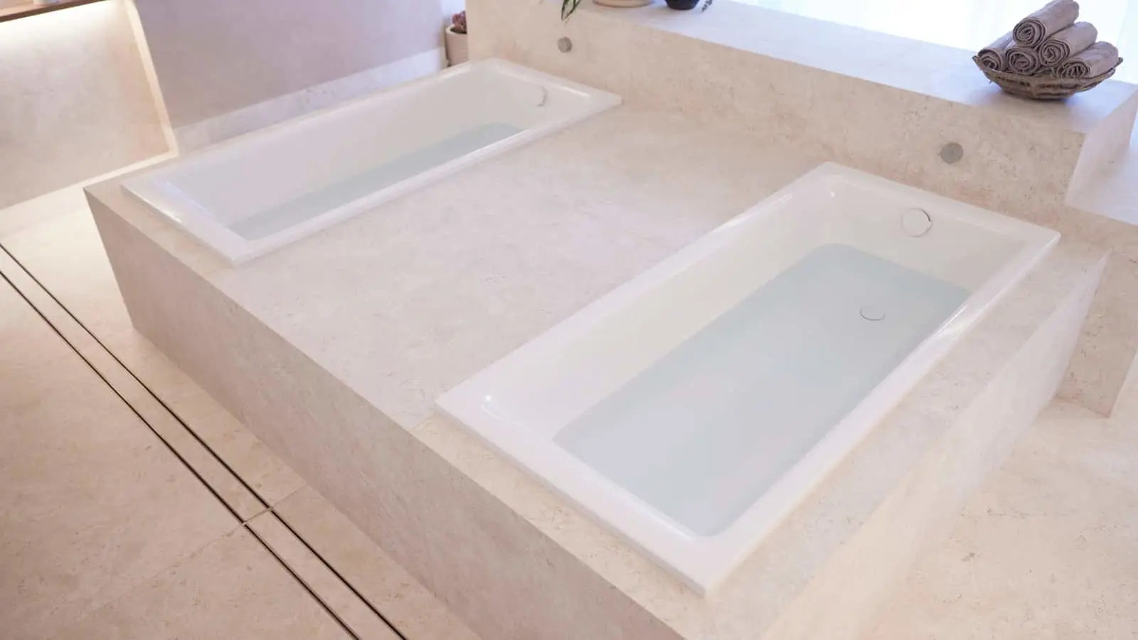 Two rectangular white bathtubs with water filled, set in cream marble platform with rolled towels in a basket nearby.