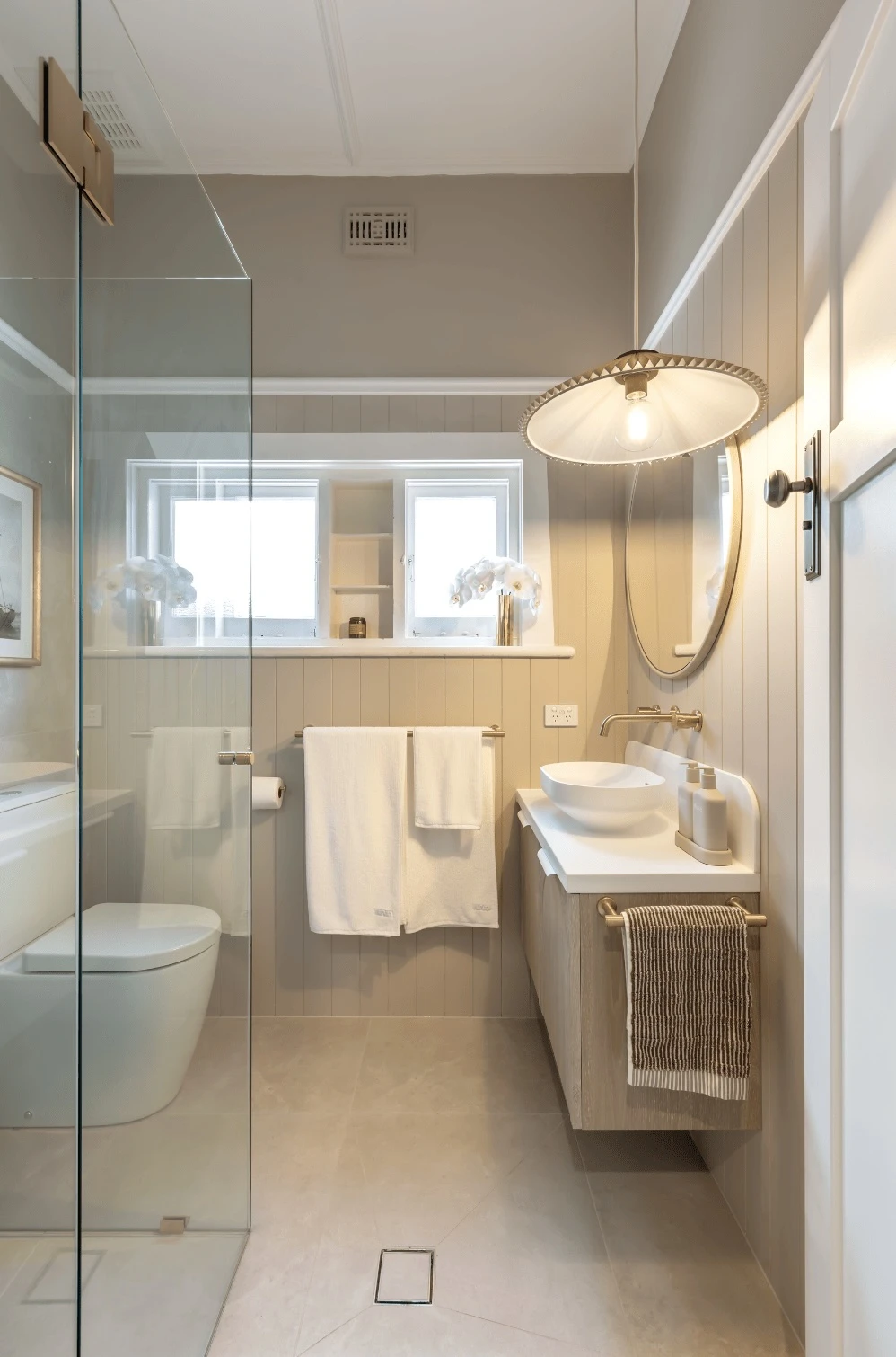 Modern bathroom with glass shower, white vessel sink, round mirror, pendant light, and beige paneled walls.