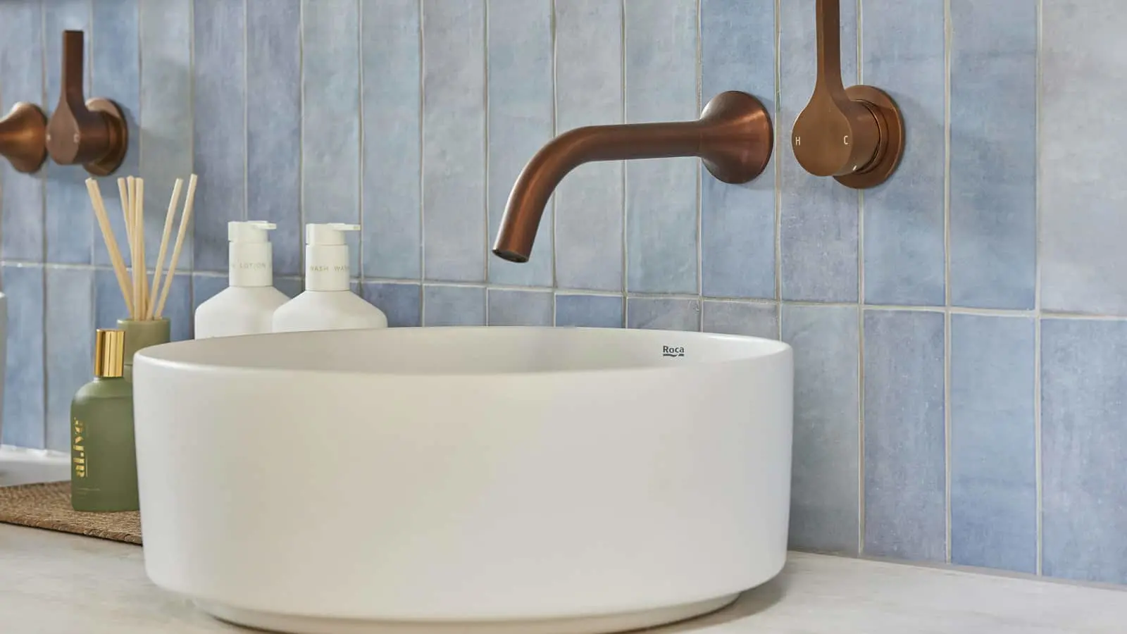 Modern bathroom sink with copper wall-mounted faucet, blue tile backsplash, and toiletry bottles on counter.