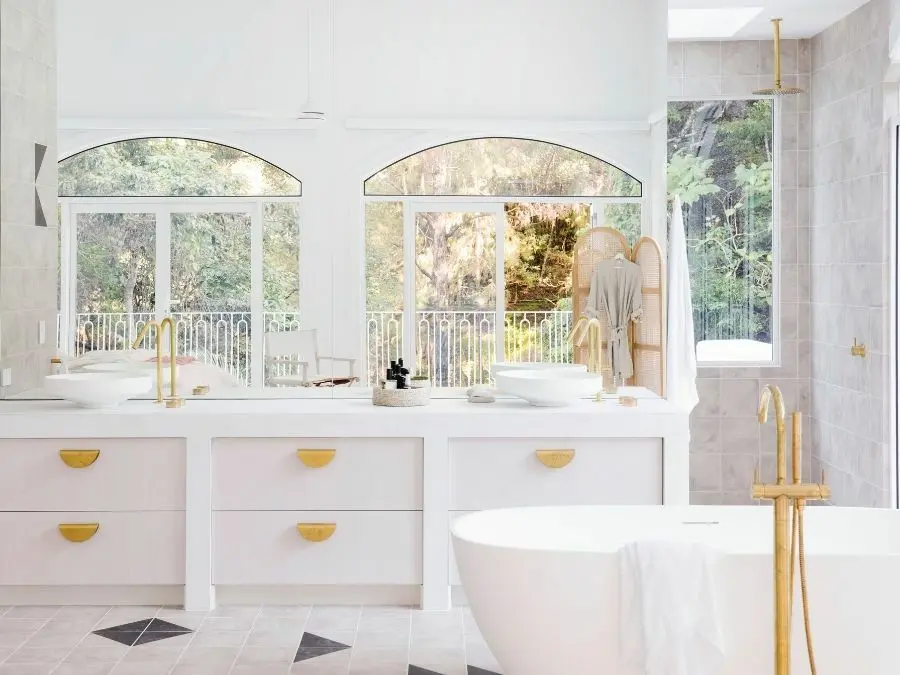 Modern white bathroom with freestanding tub, gold fixtures, and large arched windows overlooking greenery.