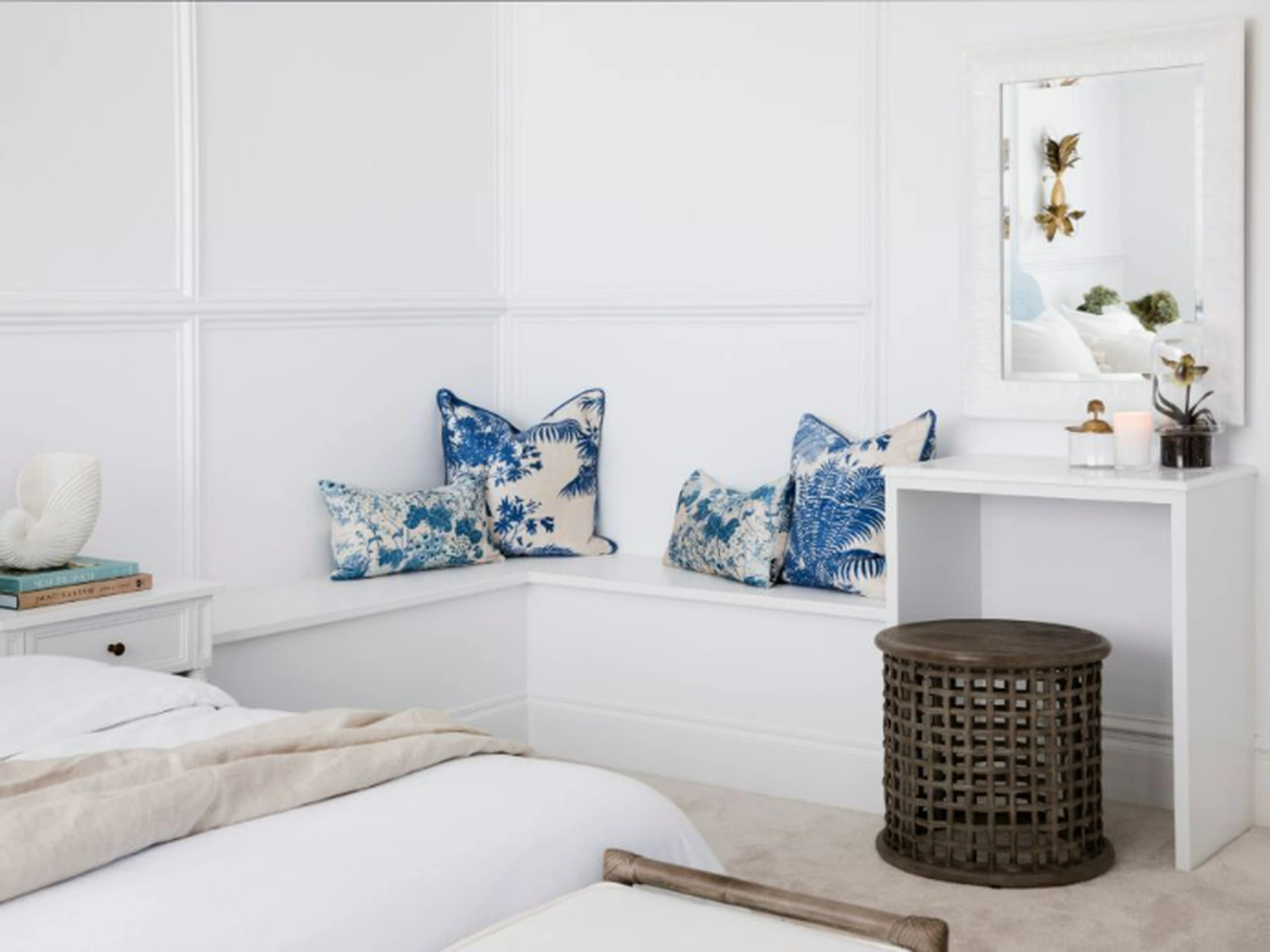 White bedroom with built-in corner bench featuring blue patterned pillows, white vanity, mirror, and wooden side table.