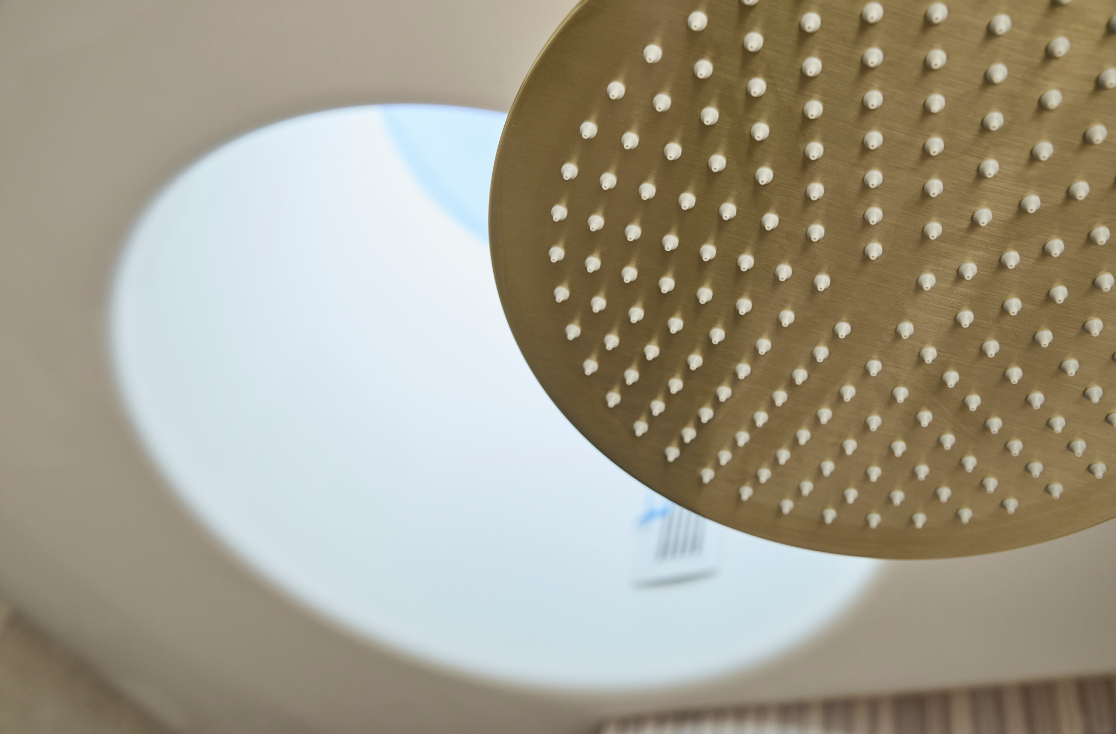 Close-up of a gold rainfall showerhead with multiple water holes, mounted on a white ceiling with a circular light fixture.