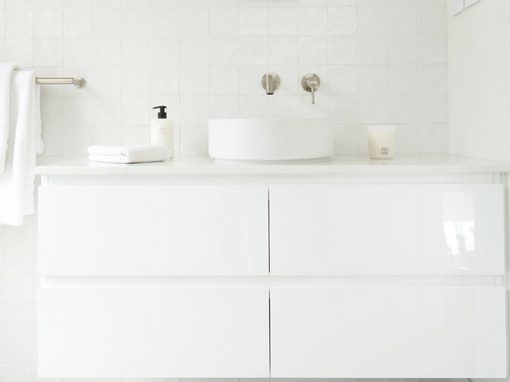 Minimalist white bathroom vanity with vessel sink, wall-mounted faucet, folded towel, soap dispenser, and candle.
