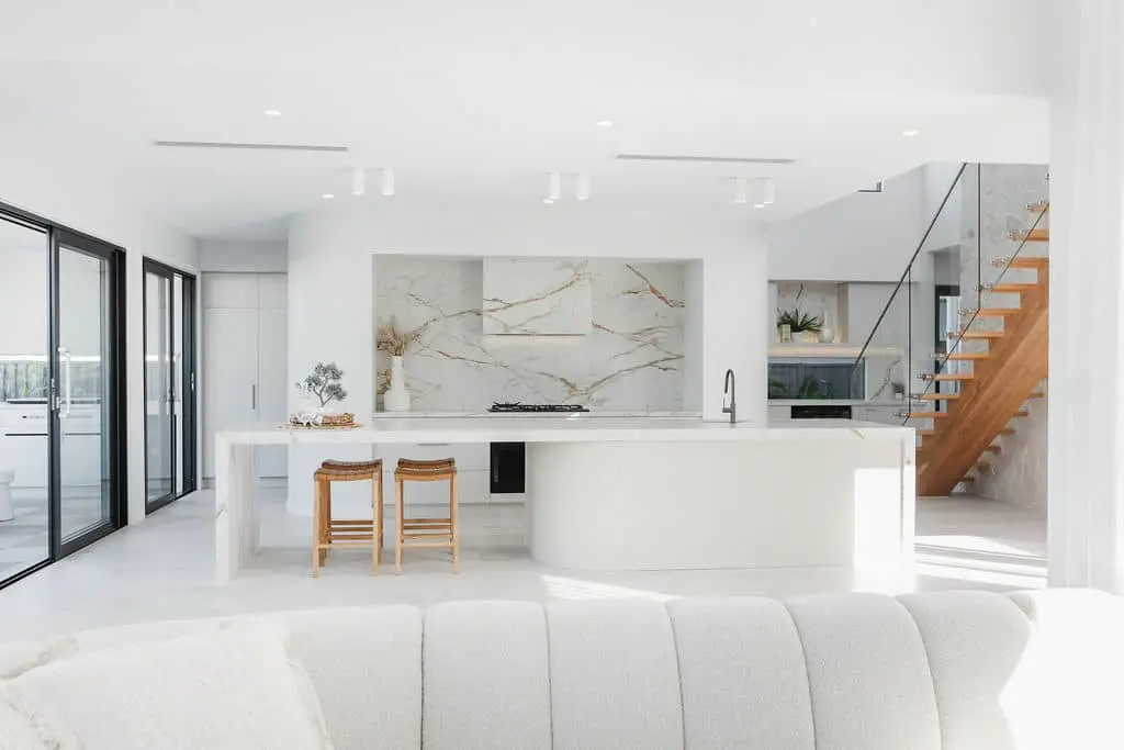 Modern white kitchen with marble backsplash, curved island, wooden bar stools, and glass staircase with wooden steps.