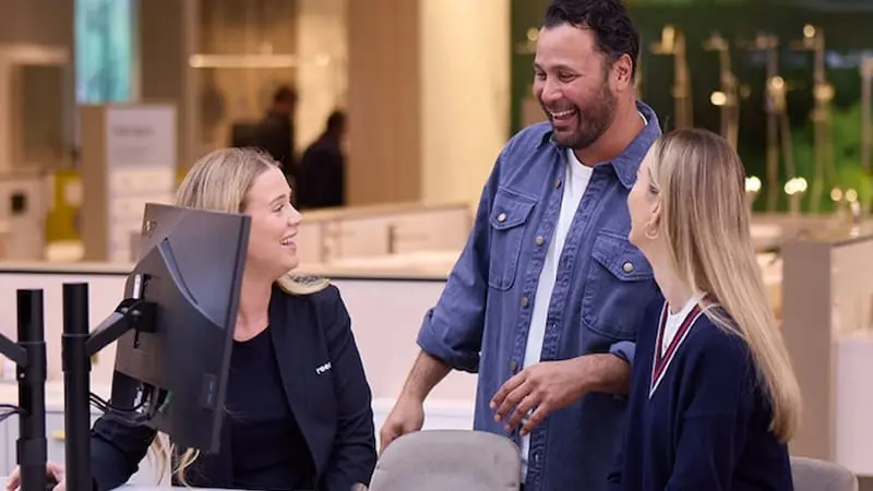Three people smiling and conversing in a retail environment with computer monitors visible in the foreground.