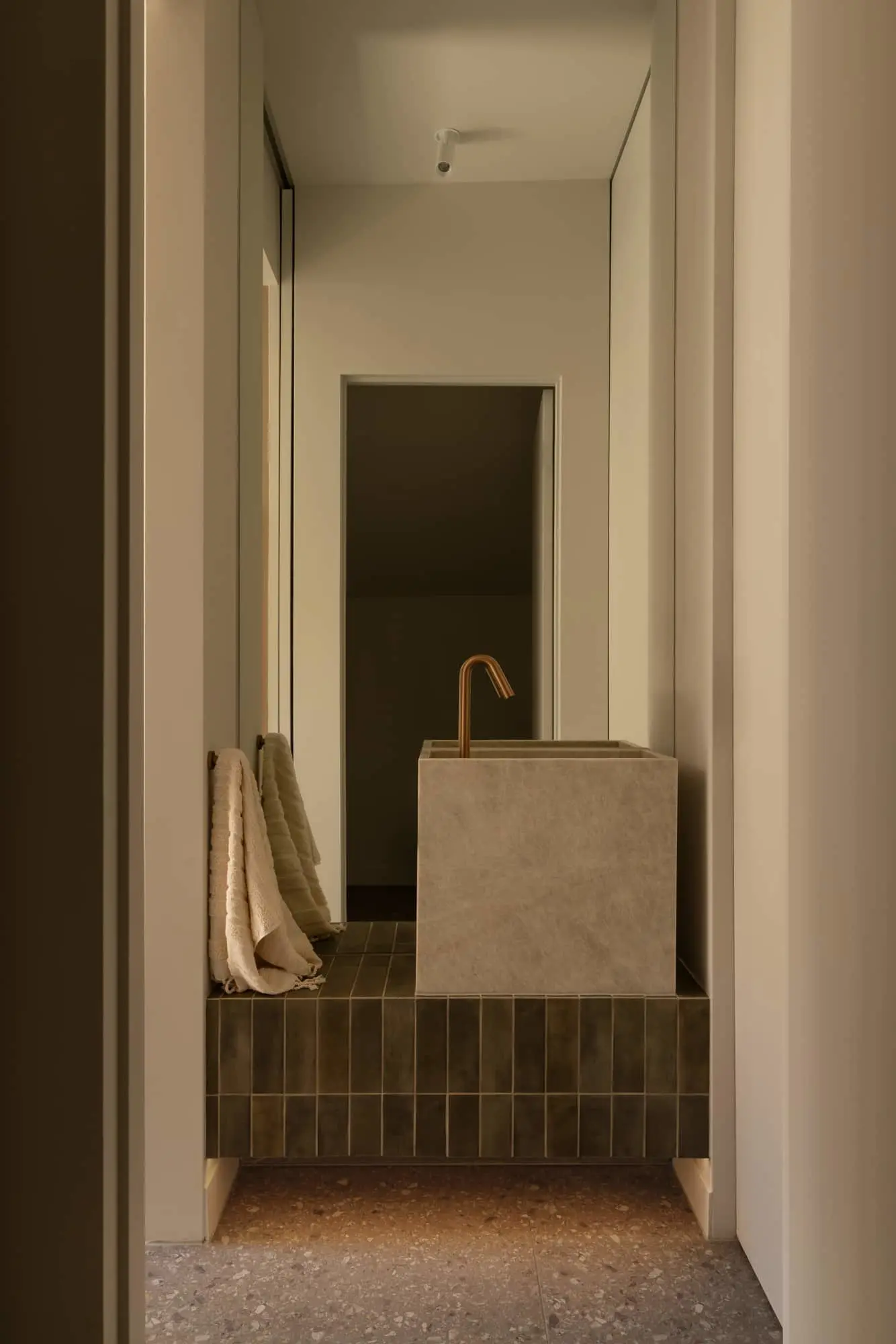 Minimalist bathroom with concrete cube sink, brass faucet, dark tiled platform, and white towels in warm lighting.