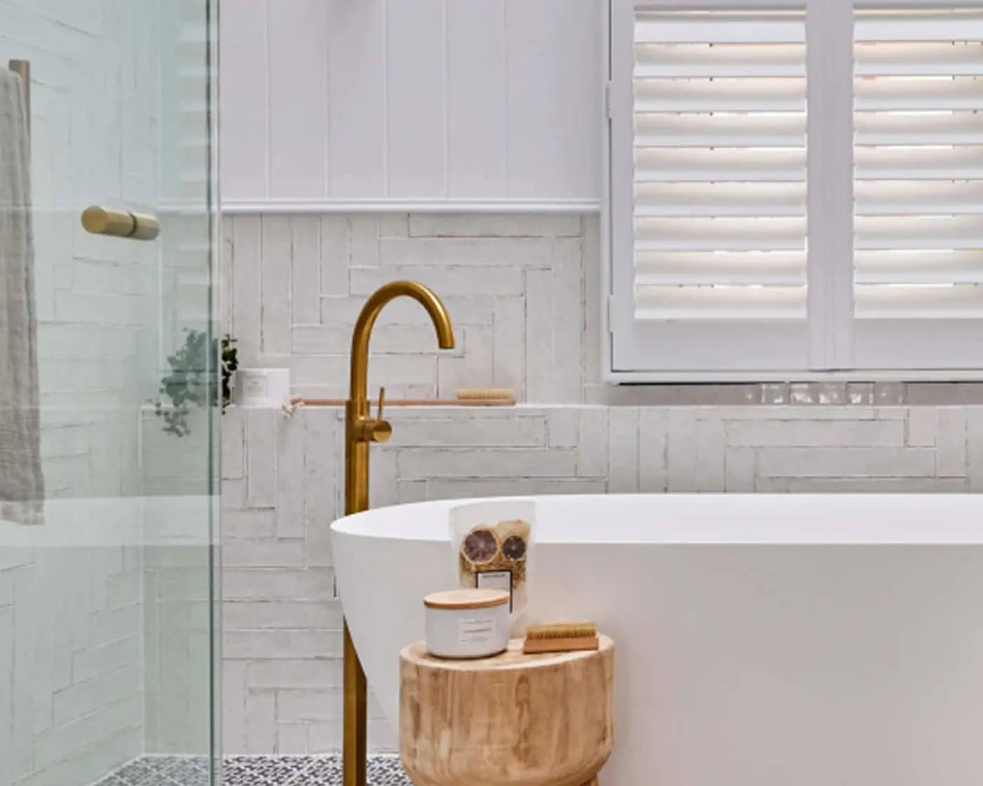 Modern white bathroom with freestanding tub, brass faucet, glass shower, wooden stool, and plantation shutters.