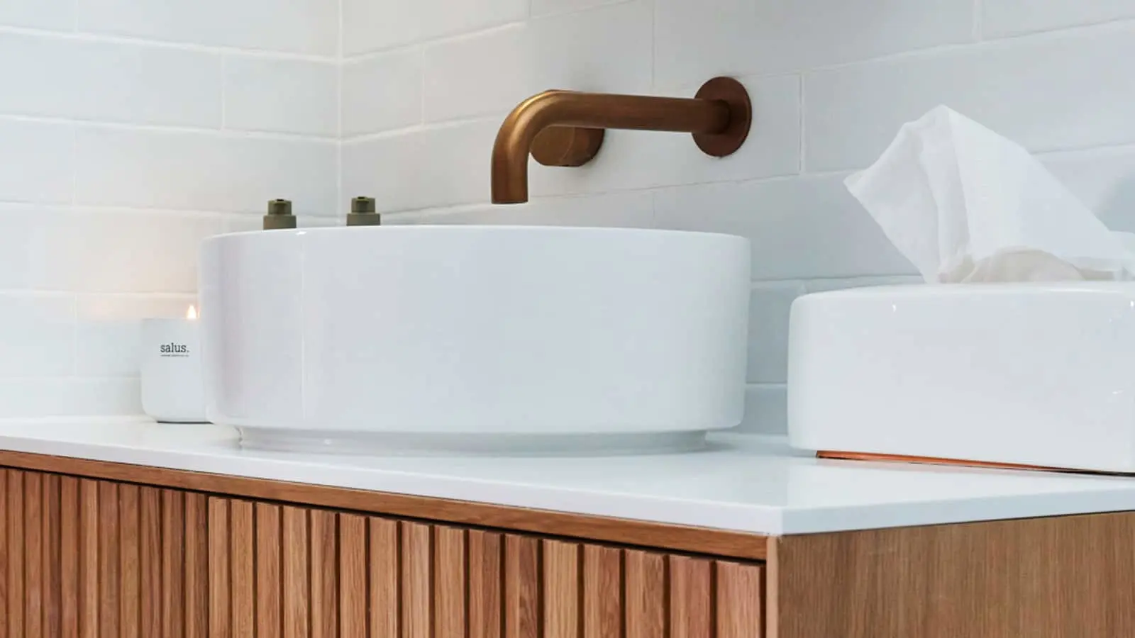 Modern bathroom with white vessel sink, bronze faucet, wooden vanity, white tile walls, and candle.