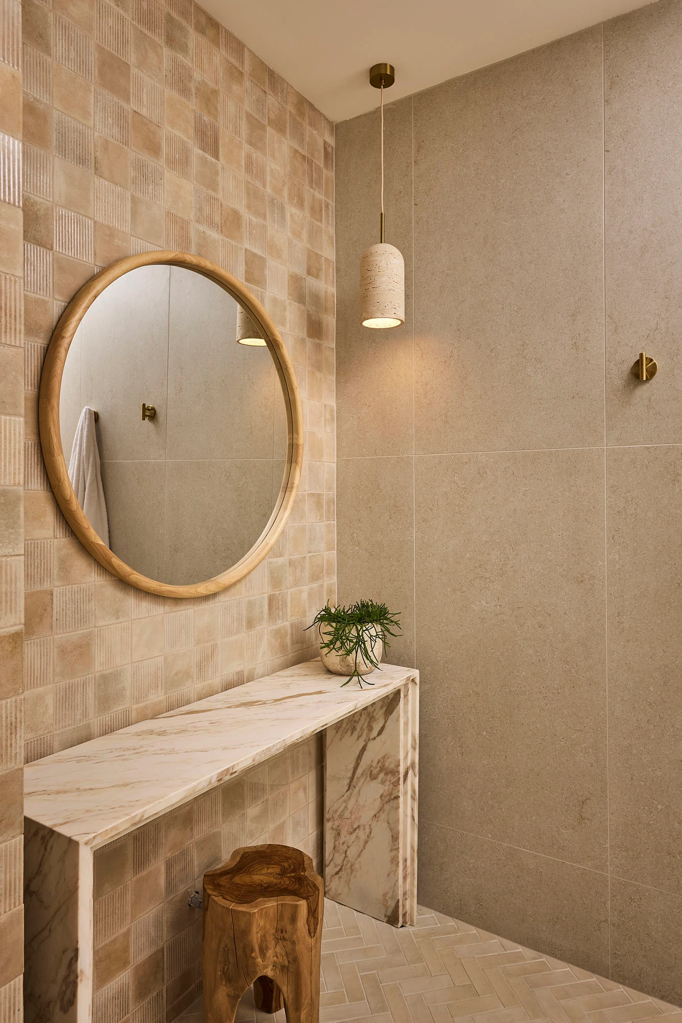 Modern bathroom with beige textured walls, round wooden mirror, marble vanity, pendant light, and small plant on counter.