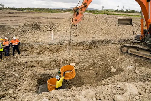 Construction workers installing orange drainage pipes in excavated ground with excavator operating nearby.