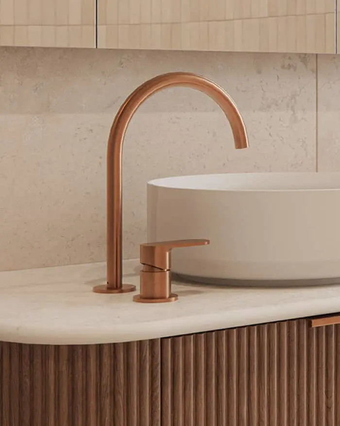 A brushed bronze arc faucet and separate handle on a stone countertop beside a white vessel sink, above a fluted wood vanity.