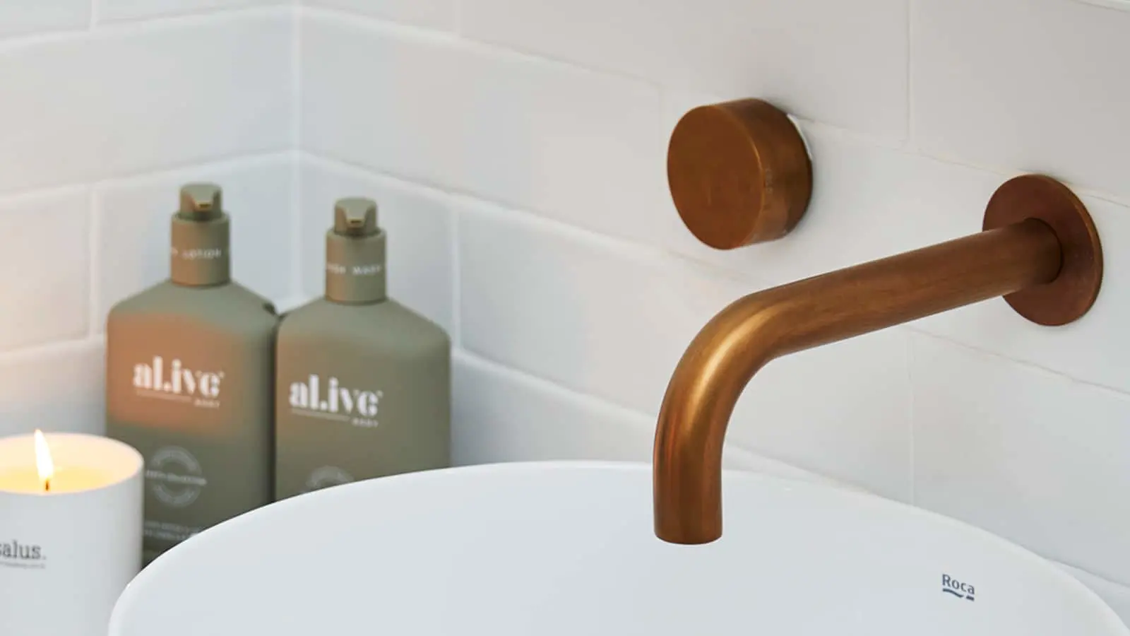 Modern bathroom sink with bronze faucet, green soap dispensers, and a lit candle against white tile wall.