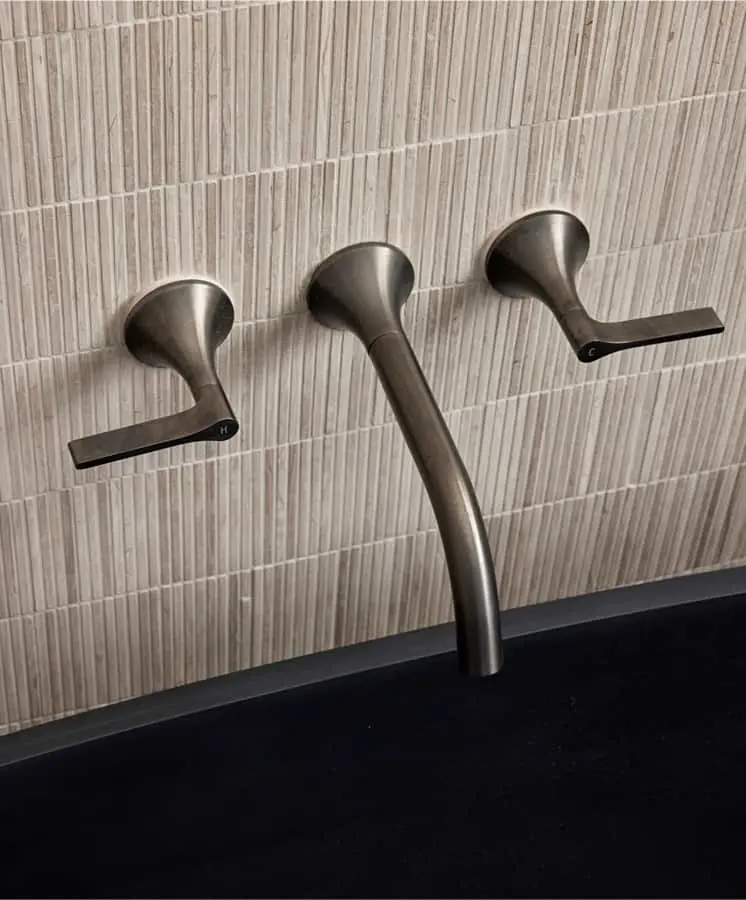 Modern wall-mounted bathroom faucet with brushed nickel finish featuring three handles against beige tiled backsplash.
