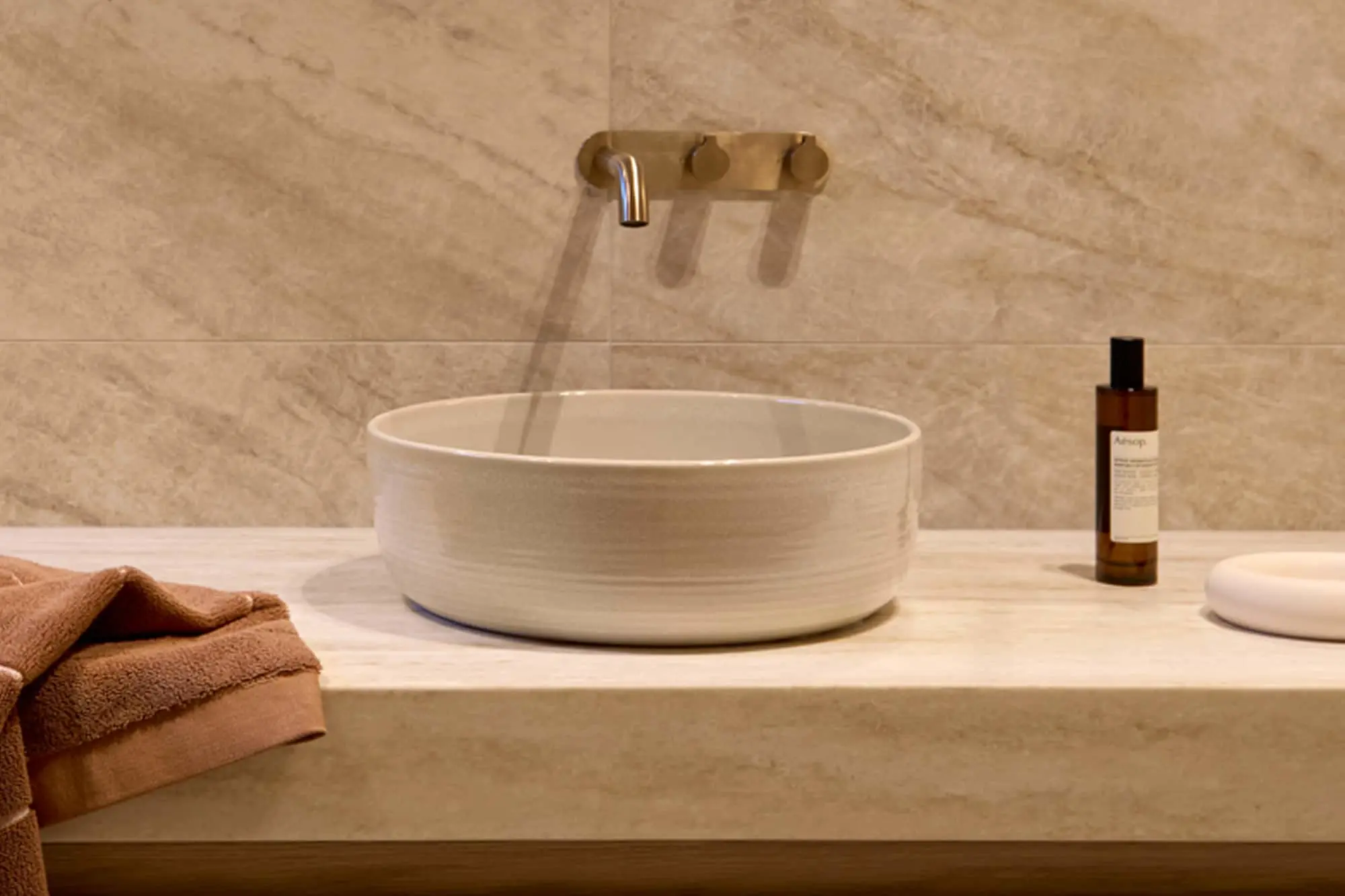 White round sink on counter with brass wall-mounted faucet, brown towel, and soap bottle against beige tile wall.