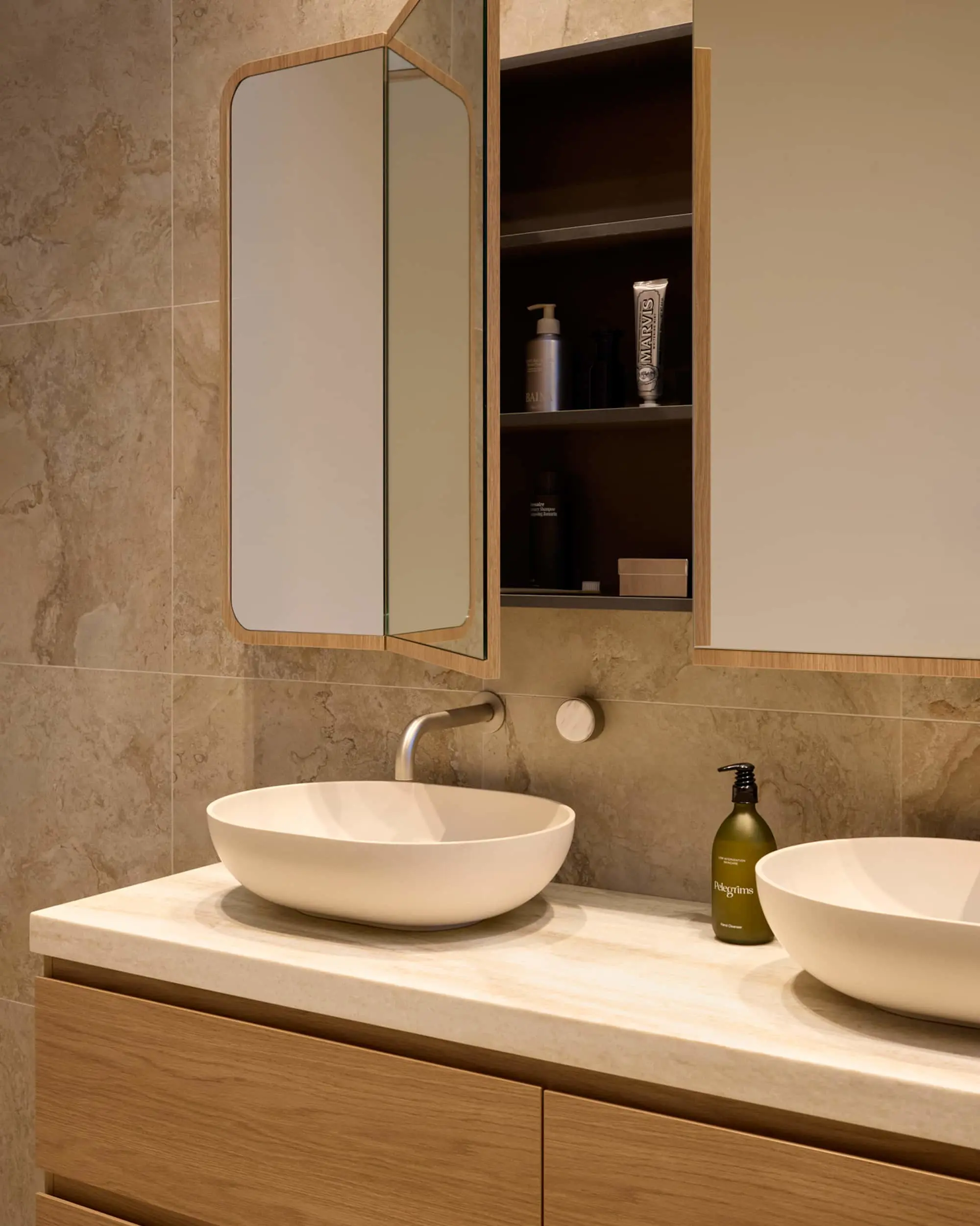 Modern bathroom with double white vessel sinks on stone counter, wood-framed mirror cabinet, and beige tile walls.