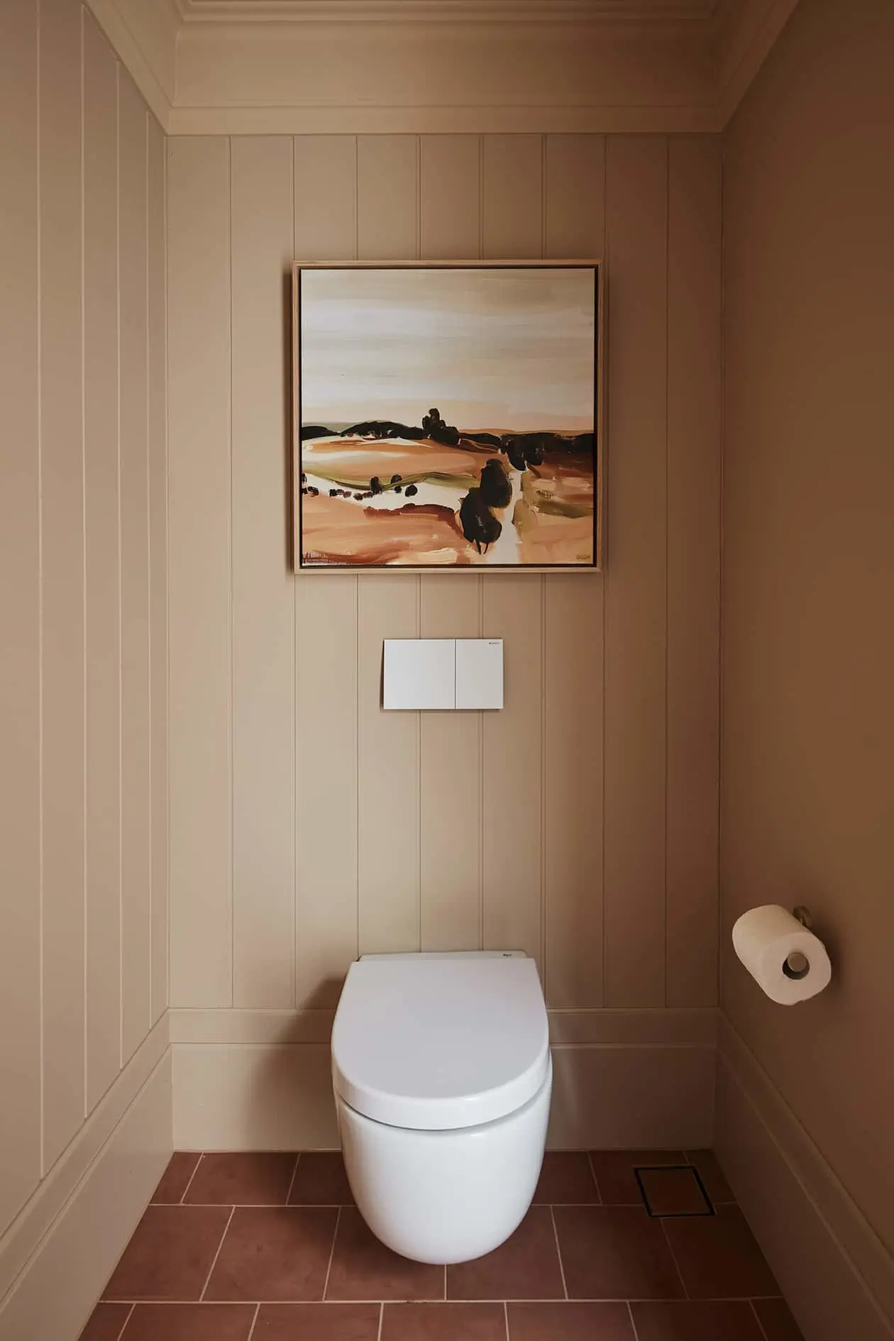 Modern bathroom with white toilet, beige paneled walls, and abstract landscape painting in warm earth tones.