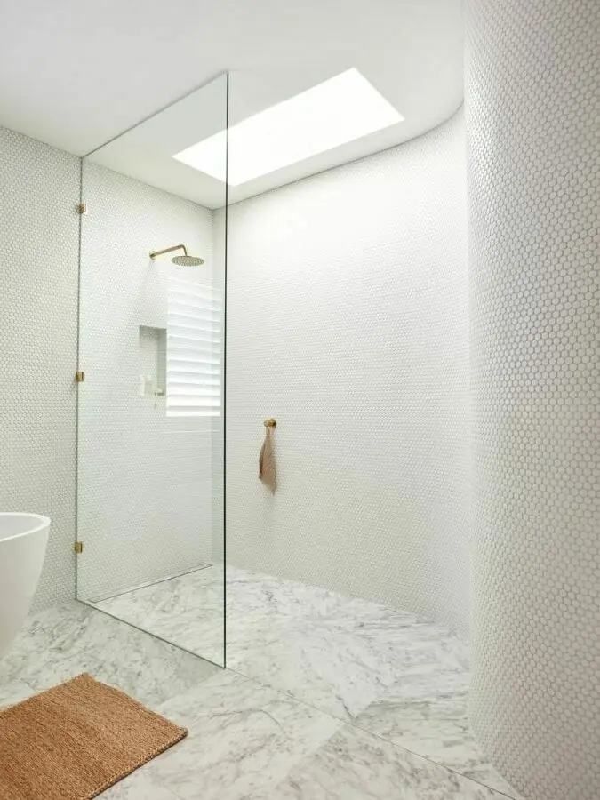 Modern white bathroom with glass shower partition, marble floor, skylight, brass fixtures, and small tan bath mat.