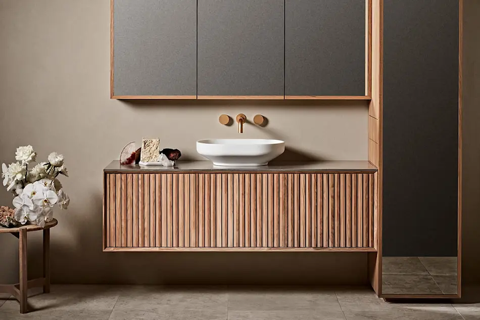 Modern bathroom with wooden vanity, white vessel sink, brass fixtures, mirrored cabinet, and white flowers on side table.