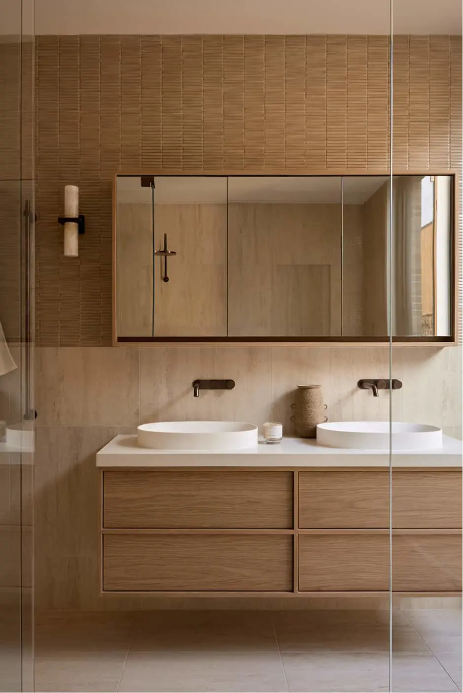 Bathroom with double vessel sinks on a floating wood vanity, large mirror, textured tan wall tiles, and bronze faucets.