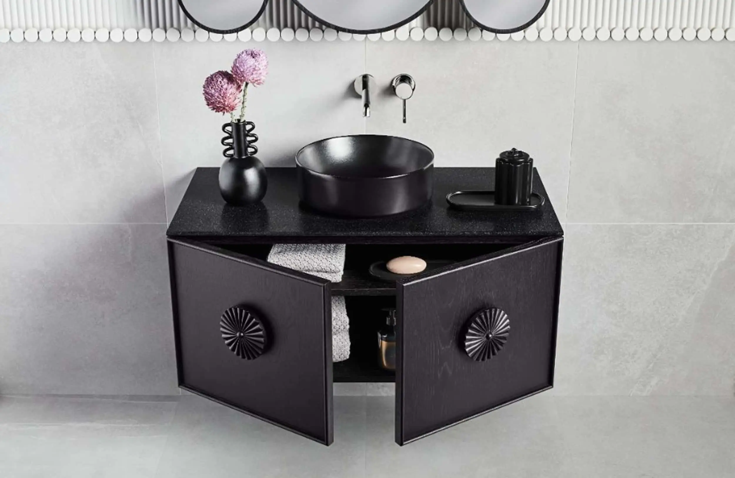 Modern black bathroom vanity with vessel sink, wall-mounted faucet, pink flowers in vase, and storage cabinet below.