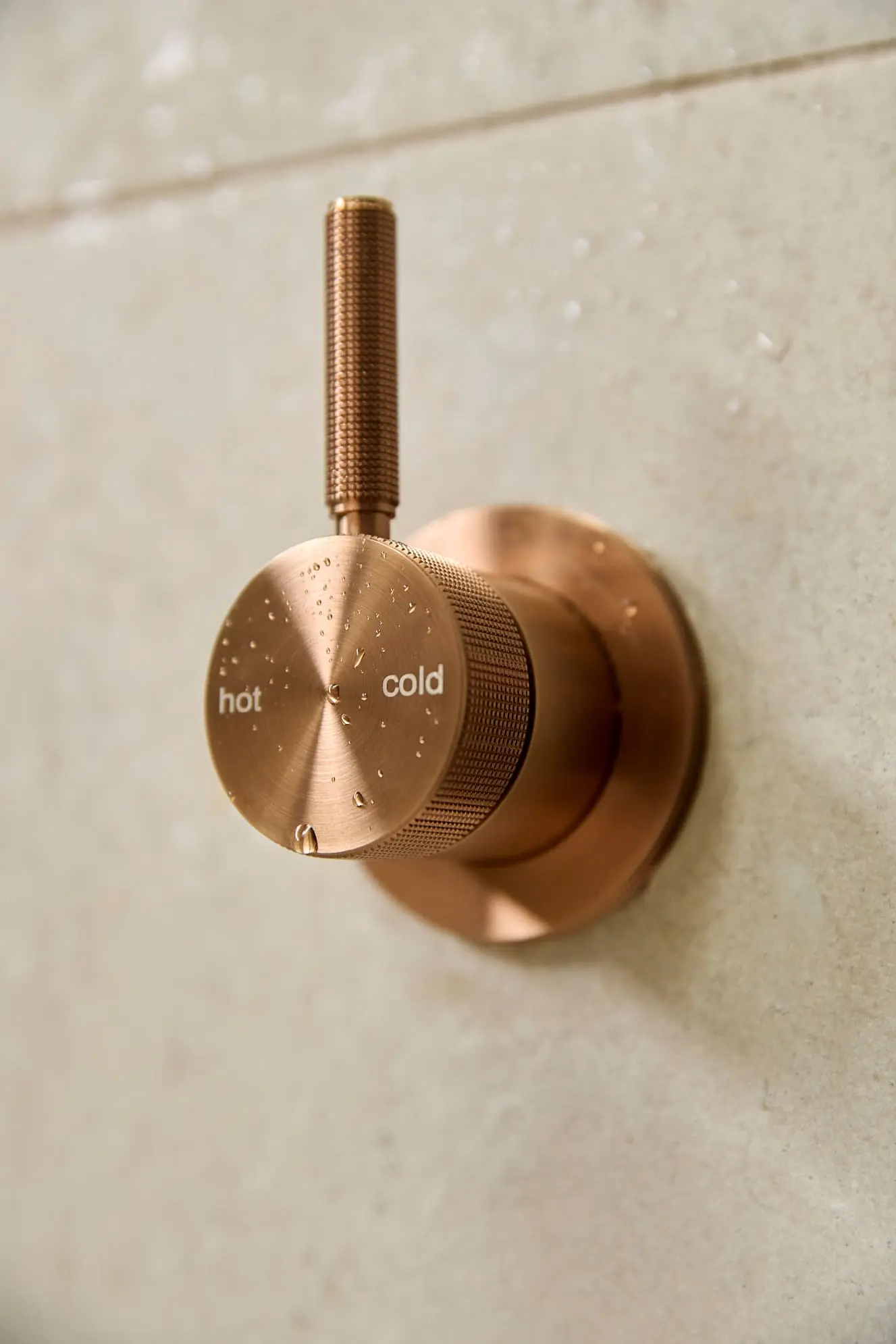 Copper shower temperature control knob with hot and cold markings mounted on a light beige wall with water droplets.
