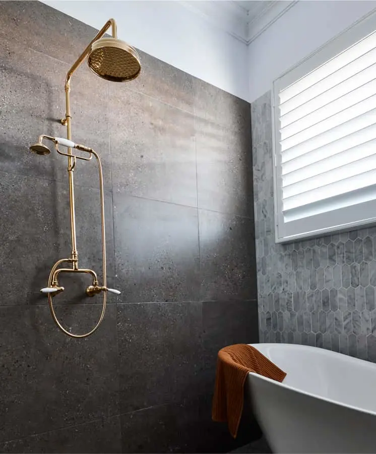 Modern bathroom with brass shower fixture on dark gray tile wall, white bathtub with orange towel, and window with shutters.