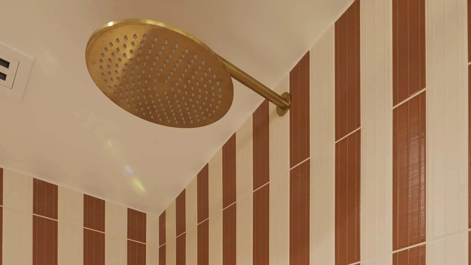 Round gold rainfall showerhead mounted on ceiling with brown and cream striped tile wall in bathroom.