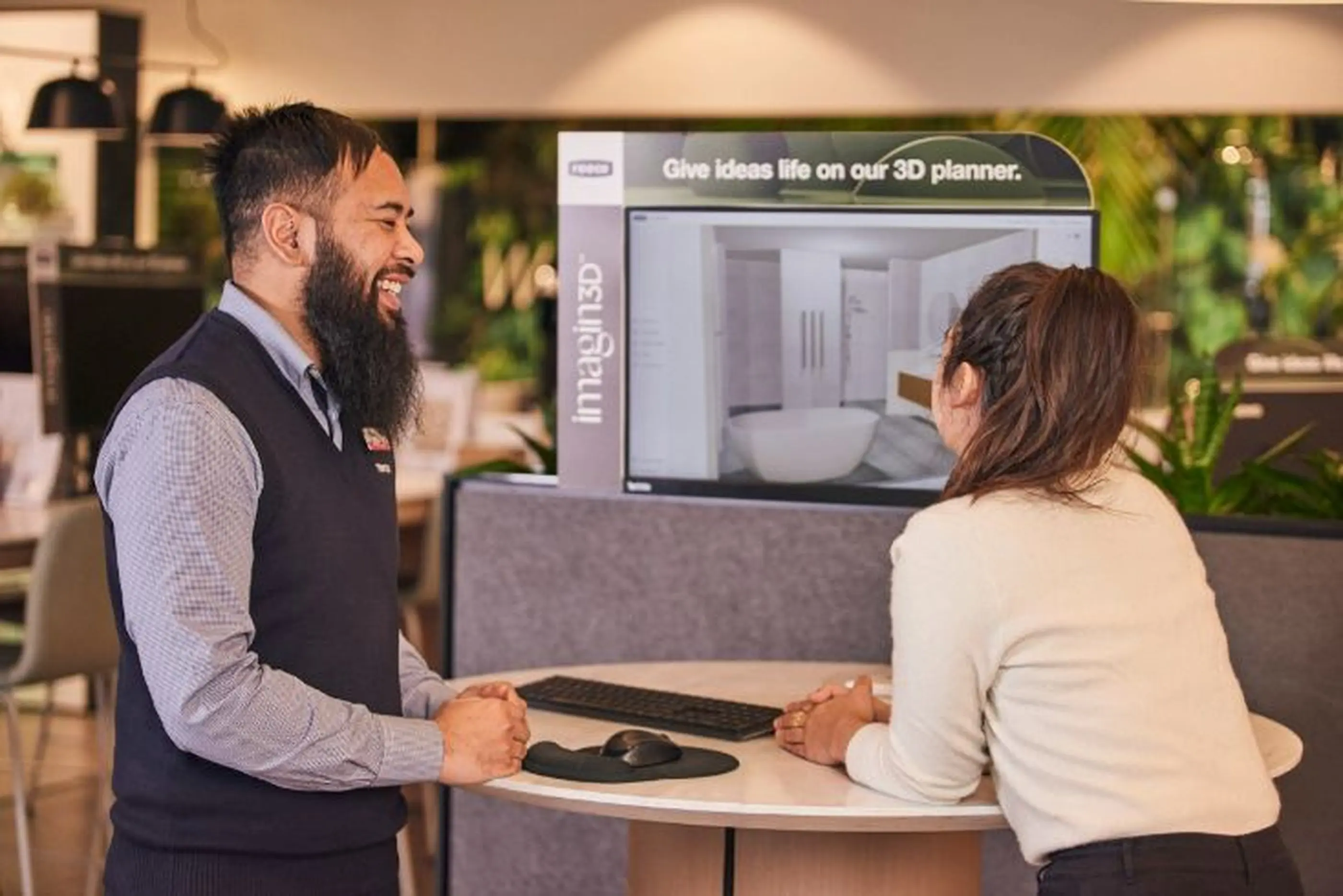Store associate helping customer use a 3D bathroom planner kiosk displaying a bathtub and sink design.