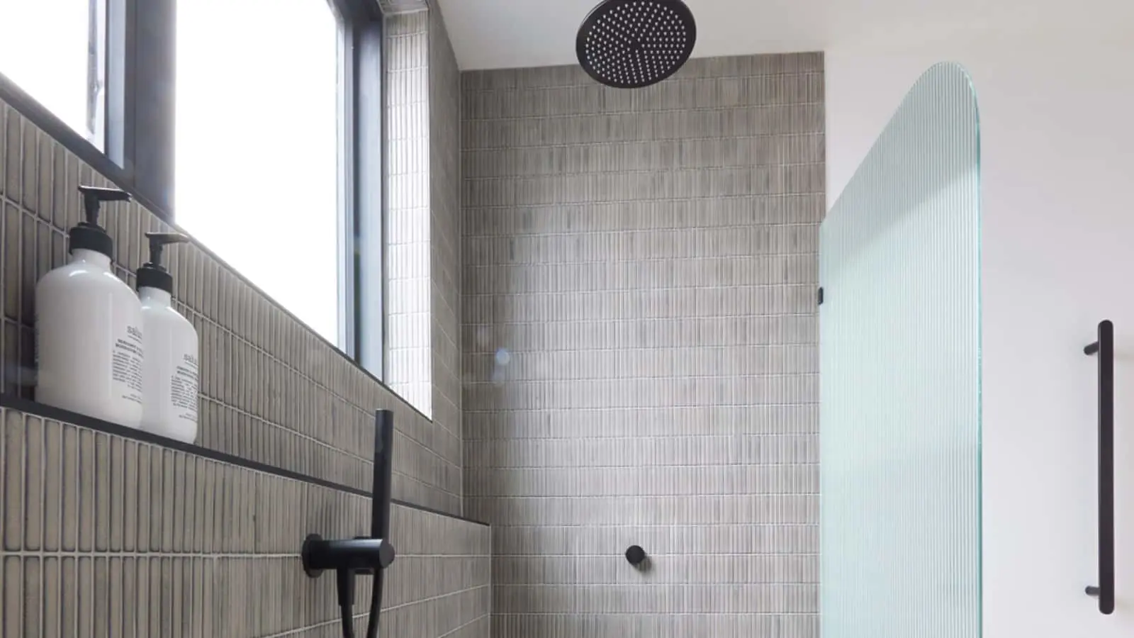 Modern shower with gray tiled walls, black rainfall showerhead, glass door, and white product bottles on shelf.