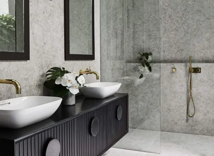 Modern bathroom with gray walls, black vanity with white vessel sinks, gold fixtures, and white orchids.