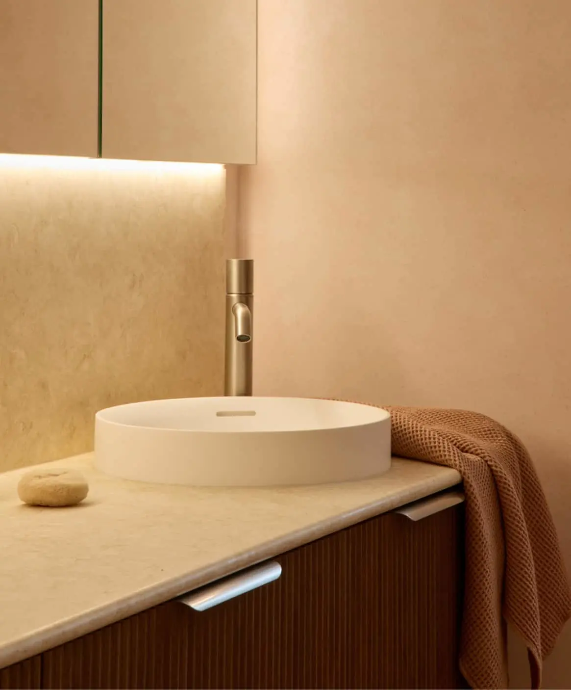 Modern bathroom with white vessel sink, brass faucet, and terracotta towel on wooden vanity with warm lighting.