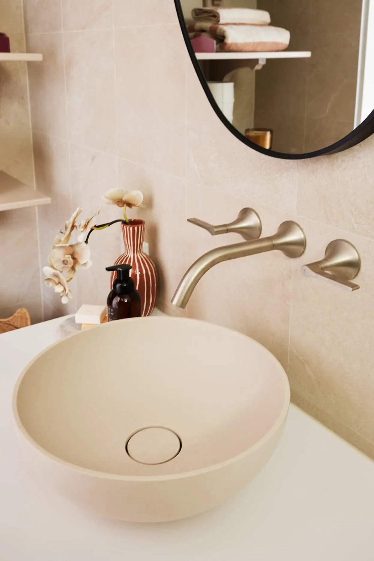 Round beige vessel sink on a white countertop with brushed gold wall-mounted faucet, round mirror, and decorative items nearby.