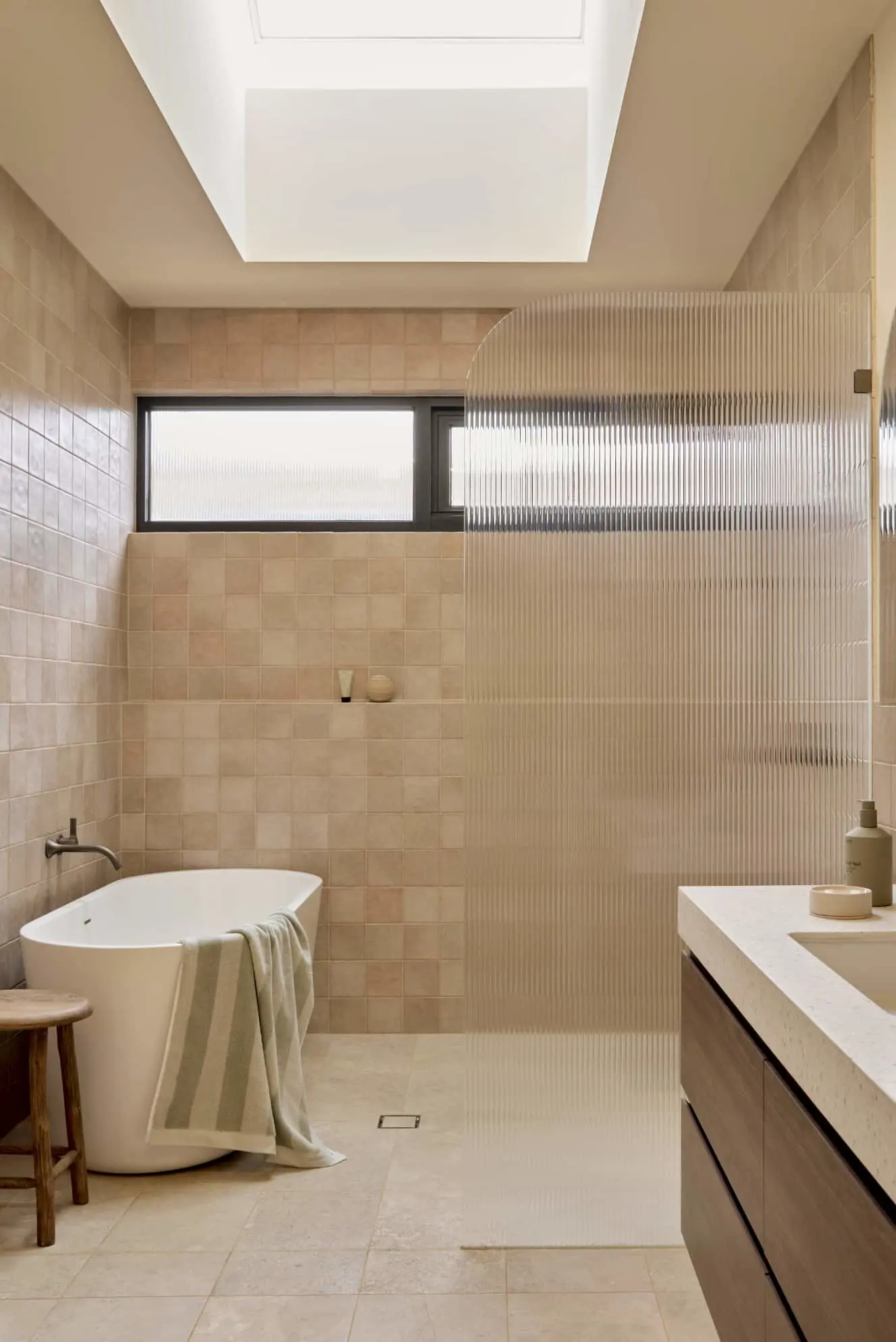 Modern bathroom with beige tiles, freestanding tub, ribbed glass shower partition, skylight, and wooden vanity.