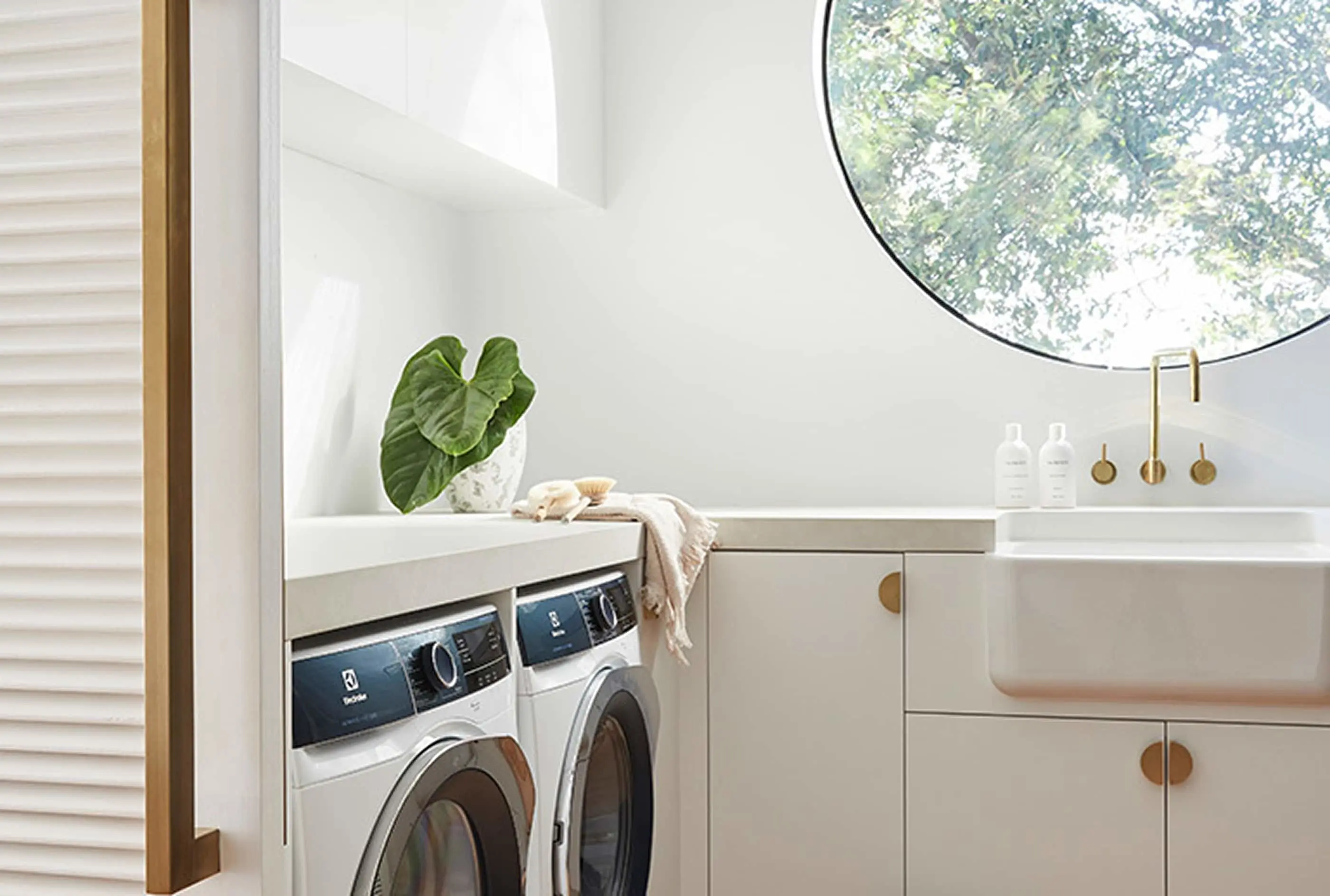 Modern laundry room with white washing machines, sink, round window showing greenery, and decorative plant leaf.