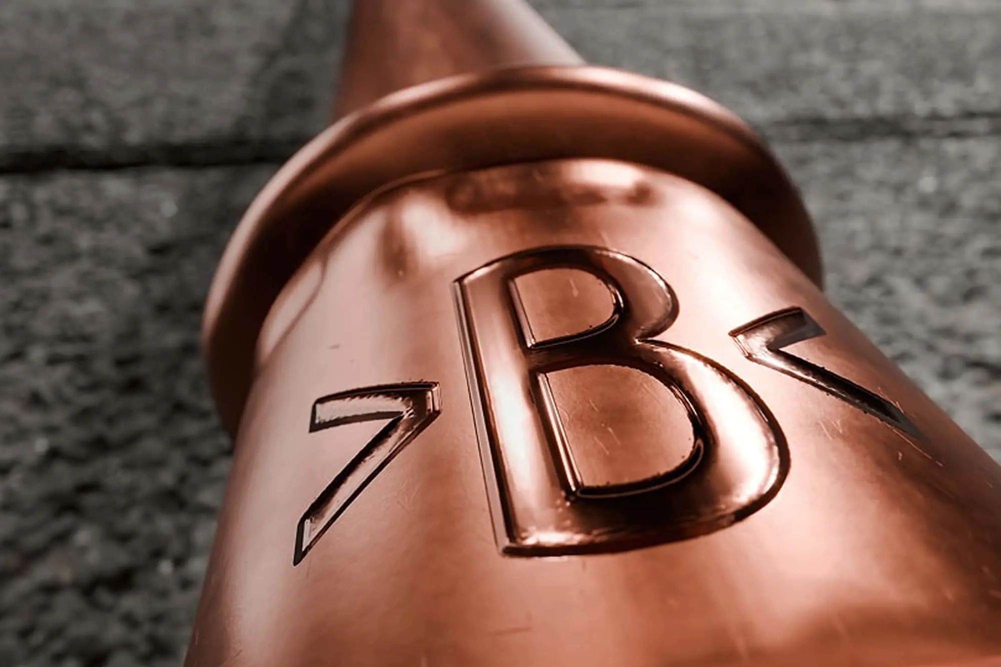 Close-up of a shiny copper pipe fitting with embossed >B< lettering against a textured gray background.