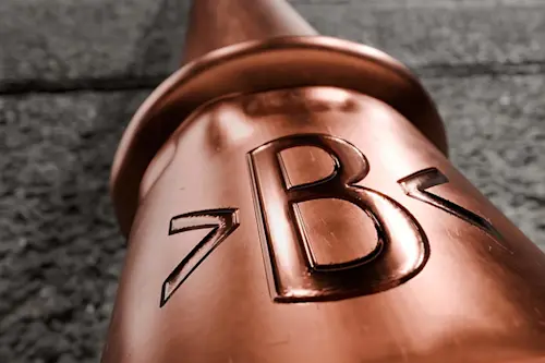 Close-up of a shiny copper pipe fitting with embossed >B< lettering against a textured gray background.