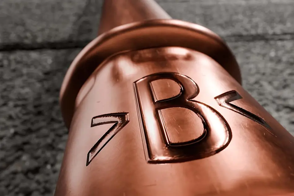 Close-up of a shiny copper pipe fitting with embossed >B< lettering against a textured gray background.