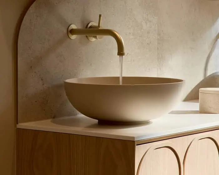 A beige bowl-shaped vessel sink on a wooden vanity with a gold wall-mounted faucet running water against a stone wall.