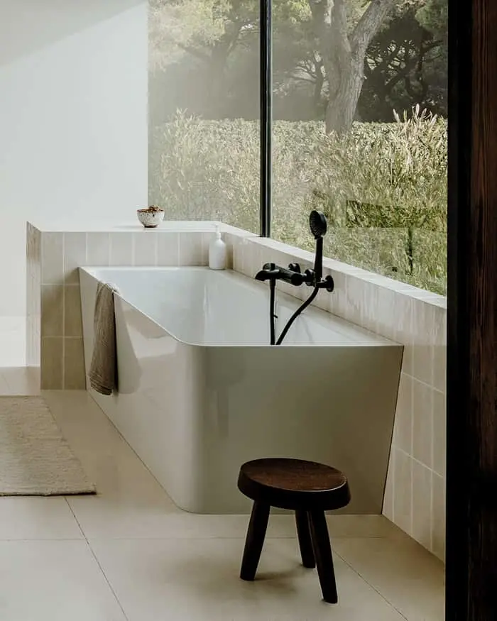 Modern white bathtub with black fixtures beside large window overlooking misty garden, wooden stool nearby.